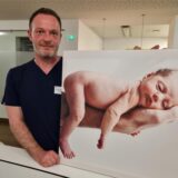 Michael Melchisedech steht im St.-Vincenz-Krankenhaus in Datteln neben einem Bild, auf dem ein Baby auf der Hand eines Mannes liegt.