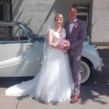 Eine Frau und ein Mann stehen in Brautkleid und Anzug, vor einem weißen Oldtimer Wagen.
