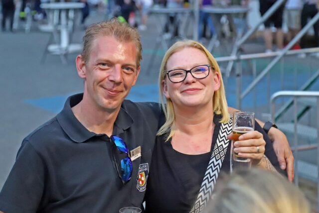 Ein Mann und eine Frau lächeln in die Kamera, im Hintergrund ist eine Menschenmenge zu sehen.