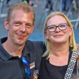 Ein Mann und eine Frau lächeln in die Kamera, im Hintergrund ist eine Menschenmenge zu sehen.