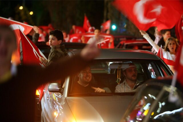 Zwei Männer sitzen in einem Auto. Auf dem Rücksitz reckt sich ein Kind aus dem Fenster und hält eine türkische Flagge. Um das Auto herum stehen jubelnde Menschen und wehen mit Türkei-Flaggen.