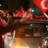 Zwei Männer sitzen in einem Auto. Auf dem Rücksitz reckt sich ein Kind aus dem Fenster und hält eine türkische Flagge. Um das Auto herum stehen jubelnde Menschen und wehen mit Türkei-Flaggen.