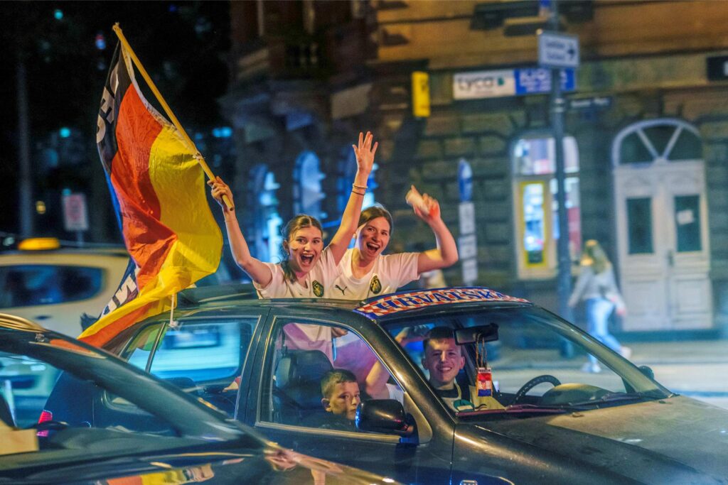 Zwei Fußballfans stehen im Auto und halten die deutsche Fußballfahne hoch. 