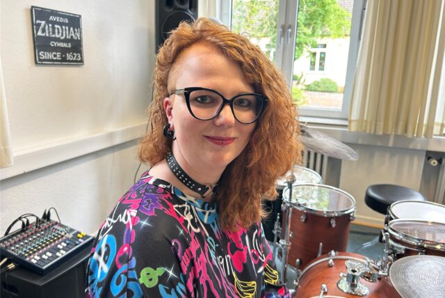 Ann-Kathrin Heitkamp hat rote, lange Locken, trägt ein buntes Shirt und sitzt an einem Schlagzeug.