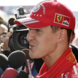 Ex-Rennfahrer Michael Schumacher spricht während eines Interviews in mehrere Mikrofone. Er trägt eine rote Jacke und eine rote Kappe.