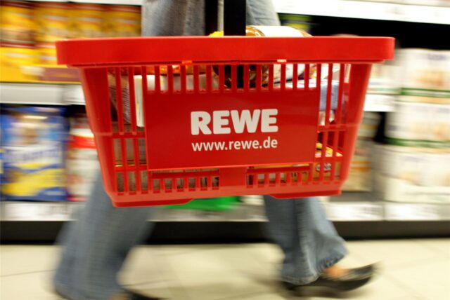 Eine Kundin mit einem tragbaren Plastikkorb mit REWE-Logo am Arm kauft ihre Lebensmittel in einem REWE-Supermarkt.