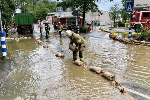 Ein Feuerwehrmann legt Sandsäcke auf eine überflutete Straße in Fröndenberg. Im Hintergrund stehen weitere Einsatzkräfte und Feuerwehrfahrzeuge