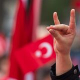 Im Vordergrund des Bildes sieht man eine Hand, die den Wolfsgruß macht. Im Hintergrund ist eine verschwommene Flagge der Türkei zu sehen.
