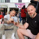 Zwei Männer sitzen in Liegestühlen auf einer Sandfläche vor dem Feuerwehrhaus der Freiwilligen Feuerwehr Heeren-Werve in Kamen und halten Cocktails in ihren Händen.