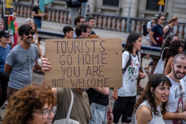 Eine Gruppe von Menschen protestiert gegen die negativen Effekte des Tourismus in Barcelona. Ein Mann hält ein Plakat hoch auf dem steht „Tourists go home, you are not welcome“.