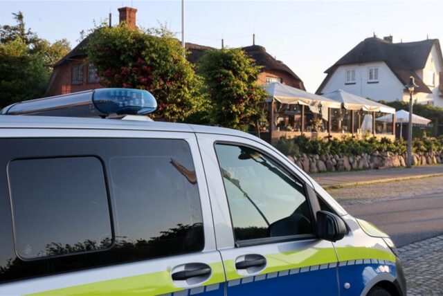 Im Vordergrund ist die Seite eines Polizeiwagens. Dieser steht am Seitenrand einer Straße an der ein Restaurant ist.