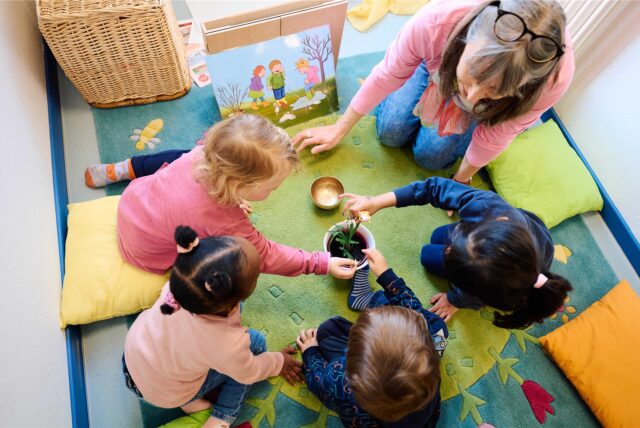 Eine Erzieherin sitzt mit vier Kindern im Kreis. In der Mitte steht eine Pflanze.