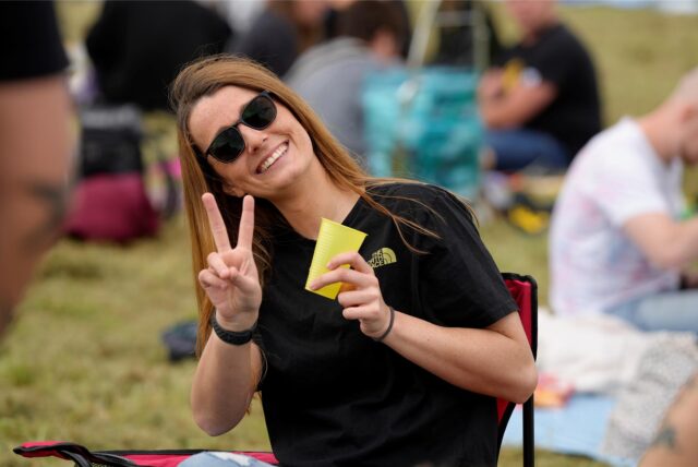 Eine Frau zeigt ein Peace-Zeichen in die Kamera. Sie trägt eine Sonnenbrille und ein schwarzes T-Shirt.