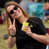 Eine Frau zeigt ein Peace-Zeichen in die Kamera. Sie trägt eine Sonnenbrille und ein schwarzes T-Shirt.