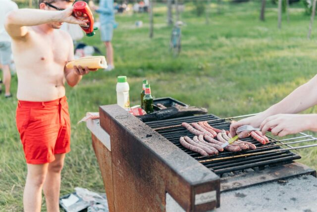 Ein Grill mit Würstchen auf einer Wiese, im Hintergrund ein Mann mit freiem Oberkörper und Baguette