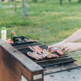 Ein Grill mit Würstchen auf einer Wiese, im Hintergrund ein Mann mit freiem Oberkörper und Baguette