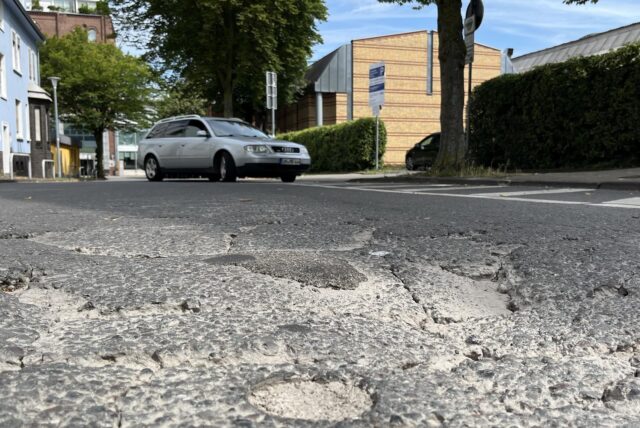 Schlaglöcher und Risse sind auf der Asphaltdecke am Westring in Unna zu sehen. Im Hintergrund fährt ein Auto auf den Parkplatz des Kinos.