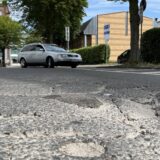 Schlaglöcher und Risse sind auf der Asphaltdecke am Westring in Unna zu sehen. Im Hintergrund fährt ein Auto auf den Parkplatz des Kinos.