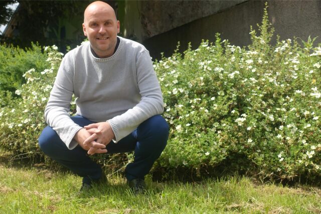 Claude Sahlmen hockt vor einem grünen Busch und lächelt in die Kamera