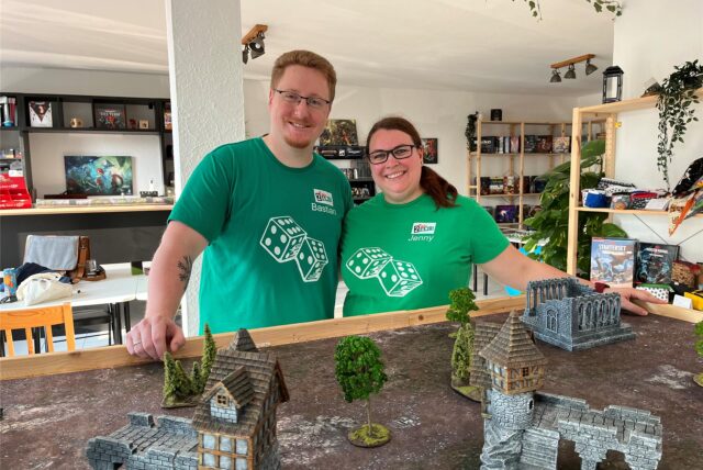 Bastian Dulczeski und Jenny Karras stehen im Laden „Two Tale Games“ in Schwerte-Ergste vor einem Tisch mit Miniatur-Häusern.