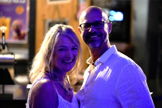 Eine Frau und ein Mann lächeln in die Kamera. Sie trägt ein weißes Top, er ein weißes Hemd und eine Brille.