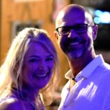 Eine Frau und ein Mann lächeln in die Kamera. Sie trägt ein weißes Top, er ein weißes Hemd und eine Brille.