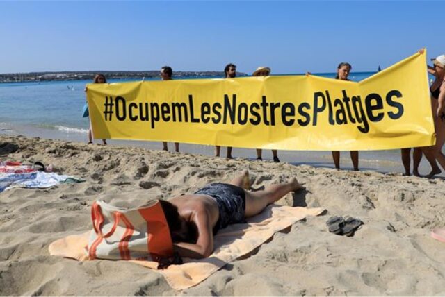 Leute halten an einem Strand auf Mallorca ein Banner „#Besetzen wir unsere Strände“ aus Protest gegen den Massentourismus auf der Balearen-Insel.
