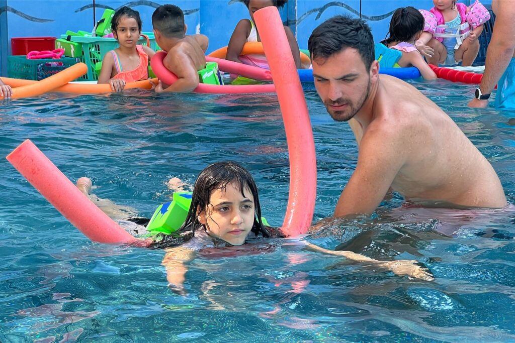 Im Bornekamp-Freibad in Unna schwimmt ein Kind mit Schwimmhilfen und Unterstützung eines Schwimmlehrers durch das Becken. Im Bornekamp-Freibad in Unna schwimmt ein Kind mit Schwimmhilfen und Unterstützung eines Schwimmlehrers durch das Becken.