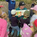 Eine Frau erklärt Kindern etwas über Lebensmittel. Die Gruppe befindet sich im Wald.