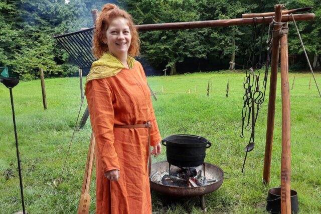 Eine junge Frau in mittelalterlicher Kleidung steht neben einem dampfenden Kessel.