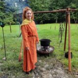 Eine junge Frau in mittelalterlicher Kleidung steht neben einem dampfenden Kessel.