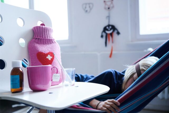 Ein Kind liegt in einer Hängematte. Davor steht ein Stuhl mit einer Tasse Tee, einer Medikamentenflasche, einem Glas und einer Wärmflasche.