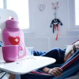 Ein Kind liegt in einer Hängematte. Davor steht ein Stuhl mit einer Tasse Tee, einer Medikamentenflasche, einem Glas und einer Wärmflasche.
