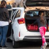 Ein Mädchen holt eine Tasche aus dem Kofferraum eines weißen Autos. Eine Frau wartet links daneben an der offenen Wagentür.