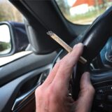 Ein Mann sitzt mit einem Joint zwischen den Fingern am Steuer eines Autos.
