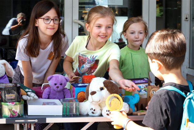 Drei Mädchen stehen auf einem Flohmarkt hinter einem Verkaufstisch und kassieren das Geld eines Jungen für ein kleines Spielzeug