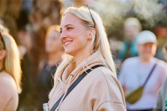 Eine blonde Frau mit einem beigen Pullover lächelt.