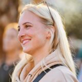 Eine blonde Frau mit einem beigen Pullover lächelt.