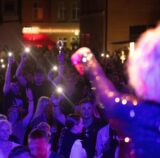 Eine Musikerin mit glitzerndem Kostüm winkt in eine Menge aus tanzenden Menschen.