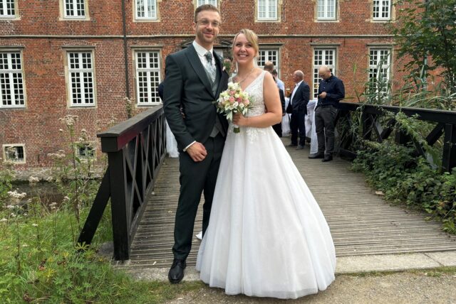 Vor dem Schloss Herten steht das Brautpaar Sobol. Kevin Sobol steht links in einem schwarzen Anzug und dunkler Krawatte. Joanna Sobol steht rechts neben ihm in einem weißen Brautkleid mit einem bunten Blumenbouquet in der Hand.