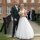 Vor dem Schloss Herten steht das Brautpaar Sobol. Kevin Sobol steht links in einem schwarzen Anzug und dunkler Krawatte. Joanna Sobol steht rechts neben ihm in einem weißen Brautkleid mit einem bunten Blumenbouquet in der Hand.