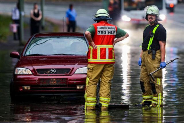 Ein Auto steckt im Hochwasser in einer Senke fest, während Feuerwehrleute die Gullydeckel öffnen, damit das Wasser nach einem Unwetter ablaufen kann.