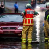 Ein Auto steckt im Hochwasser in einer Senke fest, während Feuerwehrleute die Gullydeckel öffnen, damit das Wasser nach einem Unwetter ablaufen kann.
