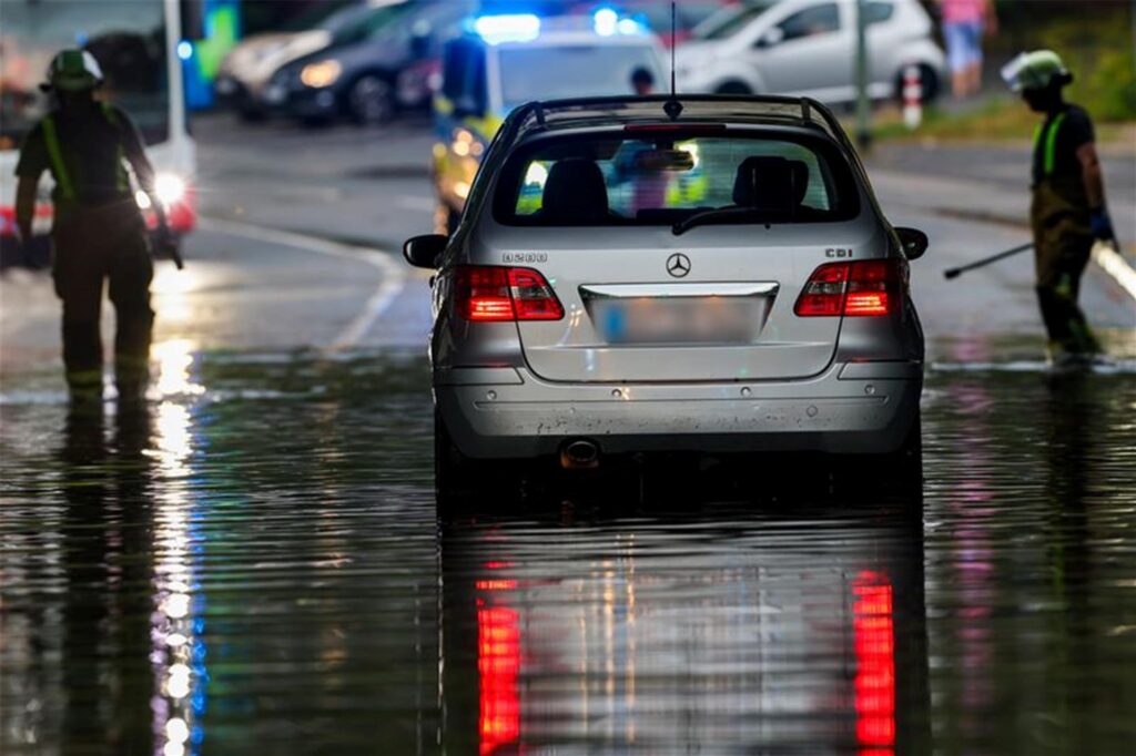 Ein Auto steckt im Hochwasser in einer Senke fest, während Feuerwehrleute die Gullydeckel öffnen, damit das Wasser nach einem Unwetter ablaufen kann. Ein Auto steckt im Hochwasser in einer Senke fest, während Feuerwehrleute die Gullydeckel öffnen, damit das Wasser nach einem Unwetter ablaufen kann.