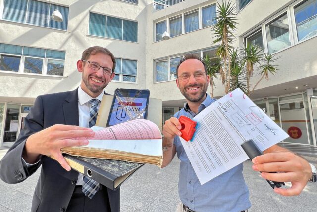 Der Erste Beigeordnete Sandro Wiggerich (l.) und Hauptamtsleiter Marc Plattfaut zeigen im Unnaer Rathaus ein Tablet sowie Papierakten mit verschiedenen Klammern.