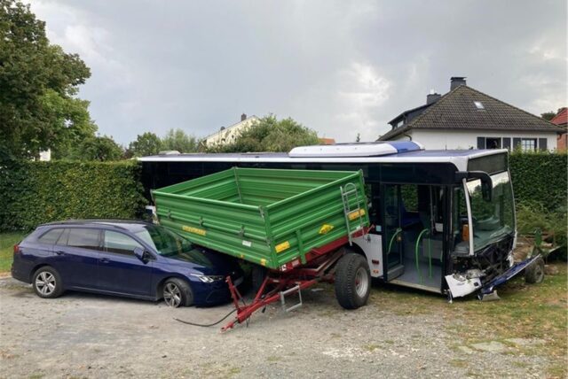 Ein Linienbus steht mit demolierter Front auf einem Hof, er hat eine Hecke durchbrochen. Daneben stehen ein Anhänger und ein Auto.