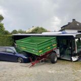 Ein Linienbus steht mit demolierter Front auf einem Hof, er hat eine Hecke durchbrochen. Daneben stehen ein Anhänger und ein Auto.