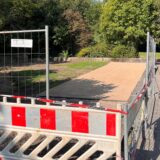 Ein Bauzaun steht vor einer hellen, rechteckigen Fläche in einem Park in Unna