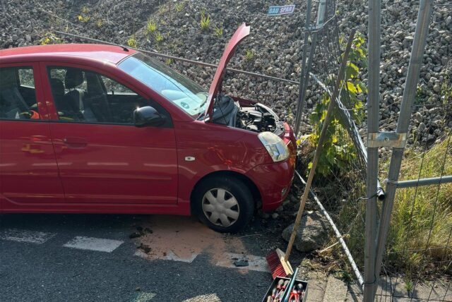 Ein Auto steht mit offener Motorhaube an einem Bauzaun.