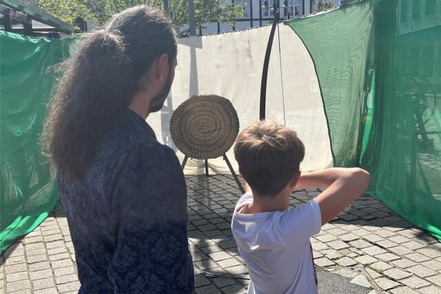 Ein Aussteller steht daneben, als ein Junge mit Pfeil und Bogen auf ein Ziel anlegt.